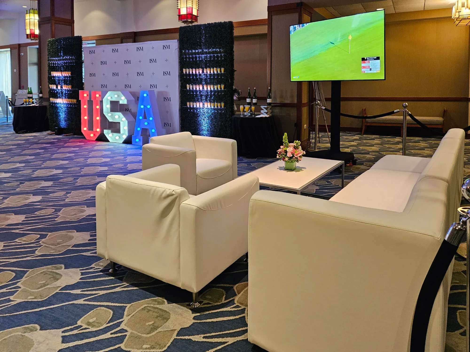 Private lounge seating in front or a USA-branded champagne wall and standing monitor displaying golf event  