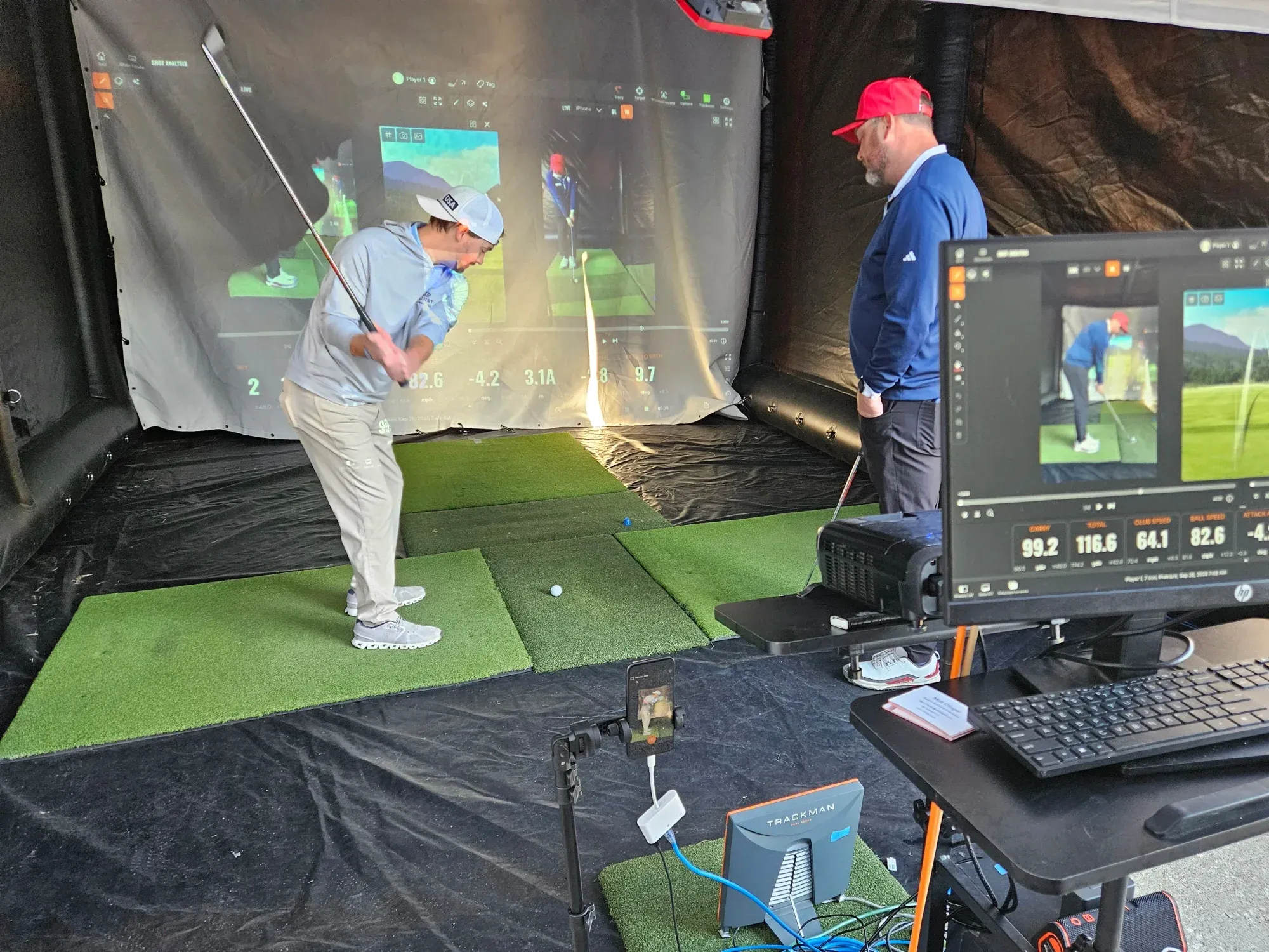 A man with a golf club in mid-swing is observed by an instructor inside a golf simulator, with computerized recording equipment in the foreground.