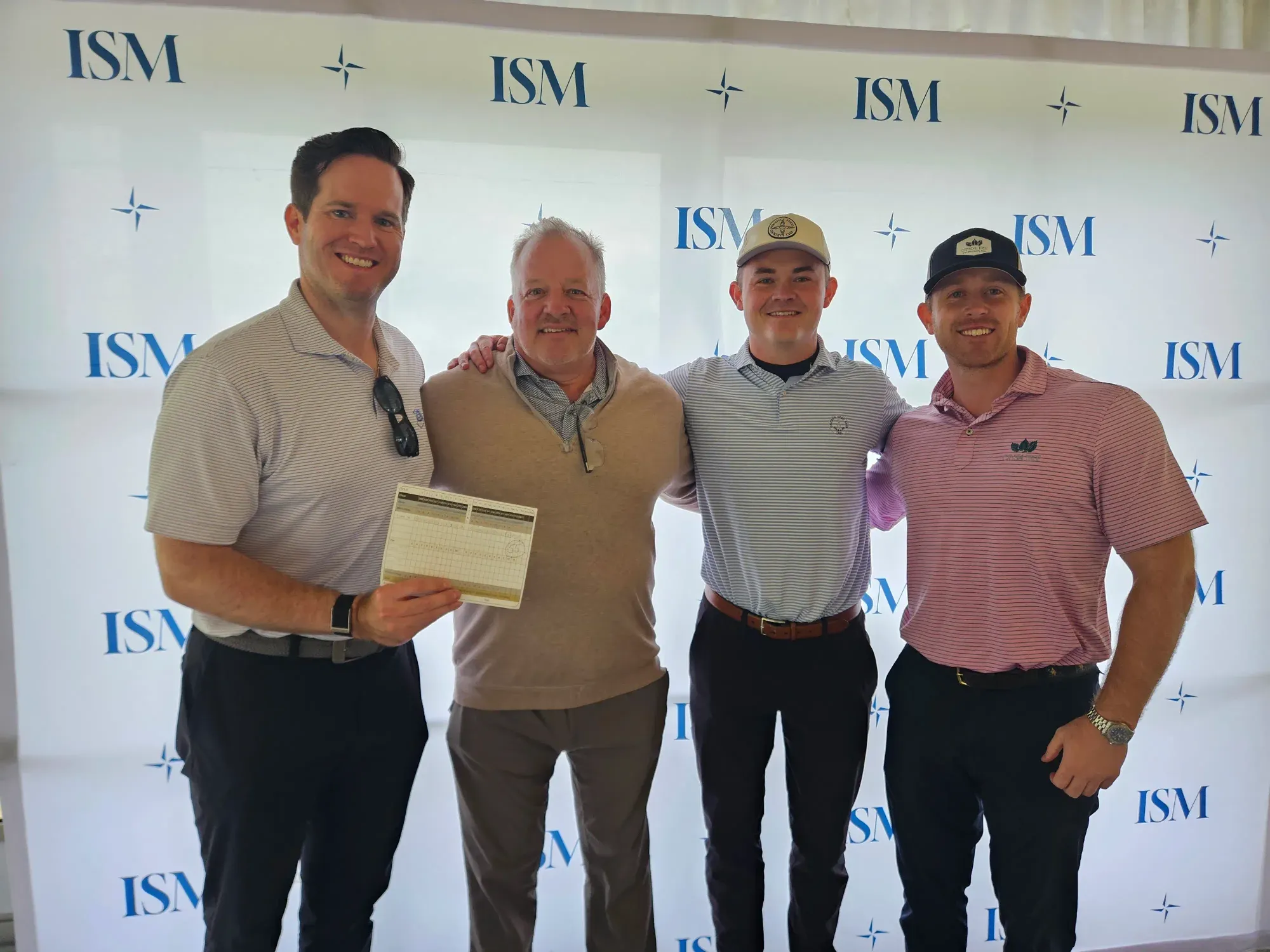 Four men in golf attire standing in front of a white ISM step-and-repeat banner. The man on the left holds their scorecard.