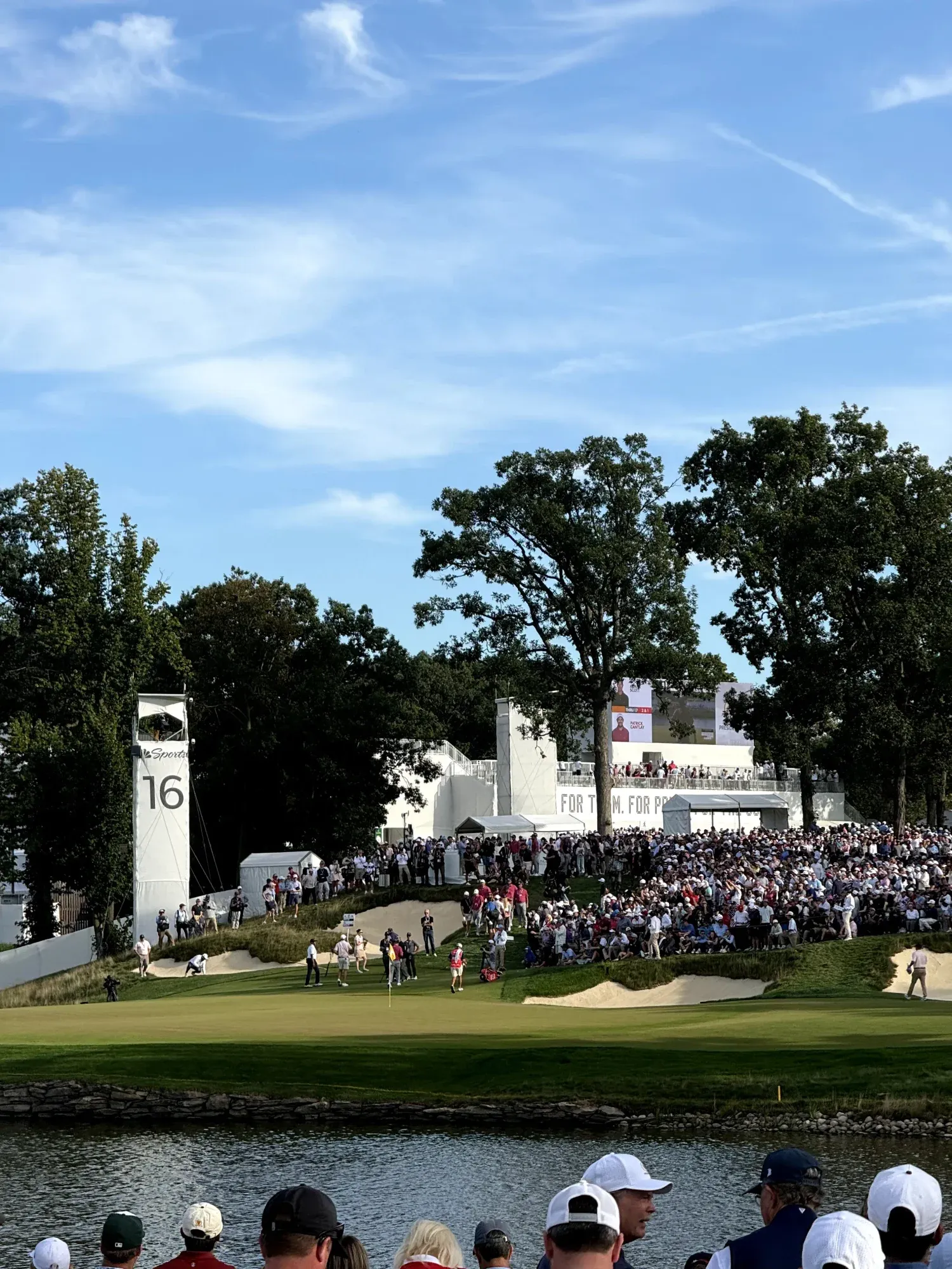Spectators visible in foreground in front of a golf water hazard. More spectators are scene near the sand bunkers on the other side.